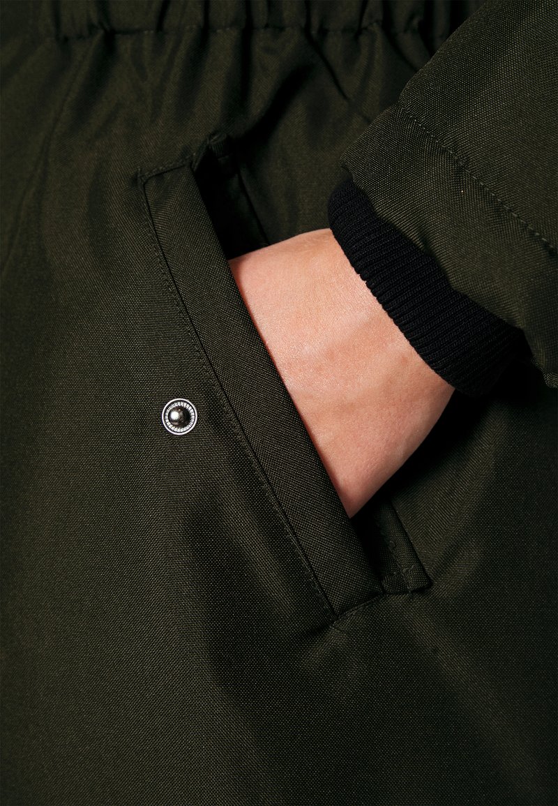 Tessuto della giacca verde oliva con texture liscia, con un primo piano di una mano in una tasca laterale e un accento di bottone a pressione argento.