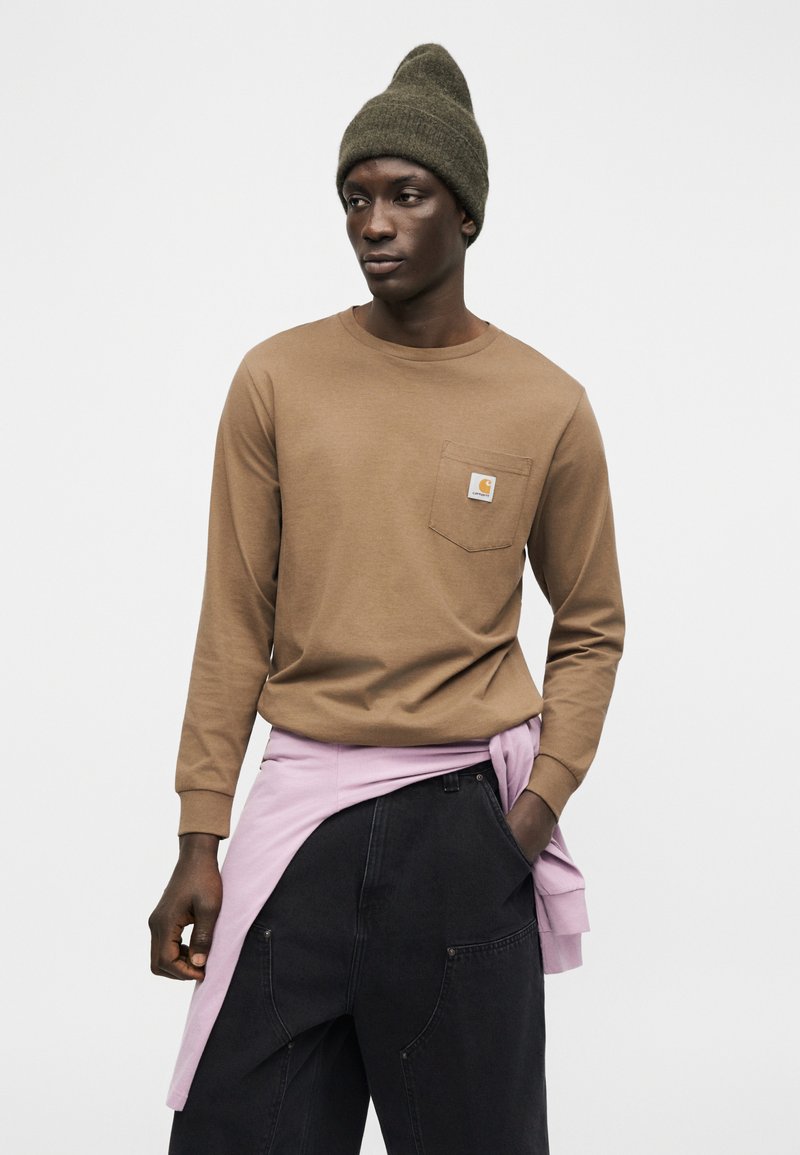 Uomo che indossa un berretto verde oliva, una camicia marrone a maniche lunghe con taschino sul petto, una felpa rosa legata in vita e pantaloni neri a gamba larga.