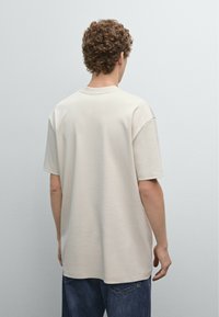 Beżowy, oversize T-shirt z krótkimi rękawami, okrągłym dekoltem i gładką bawełnianą strukturą, noszony z luźnymi dżinsami, widziany z tyłu.