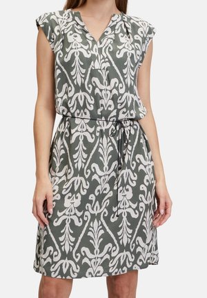 Femme portant une robe grise sans manches avec un motif floral abstrait blanc et une fine ceinture noire à la taille, debout devant un fond uni.