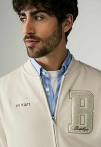 Lys beige zipjakke med broderi af "Williamsburg NY STATE", kontrasteret med en tekstureret "B" lap. Båret over en blåstribet skjorte.