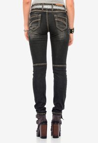 Schwarze Skinny-Jeans mit Kontrastnähten, Seitenreißverschlüssen an den Knien, Gesäßtaschen und einem gemusterten Gürtel. Getragen mit braunen, blockhohen Stiefeletten.