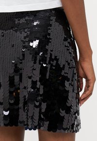 Juoda blizgučių mini sijonas su prigludusia siluetu, puošia dideli apvalūs blizgučiai, kurie atspindi šviesą, sukuriant blizgantį tekstūrą.