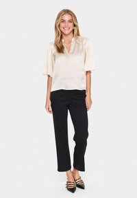 Blusa di satin beige chiaro con scollo a V e maniche a sbuffo corte, abbinata a pantaloni neri su misura e décolleté neri a punta.