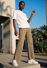 Mladá osoba v bílém tričku, béžových kalhotách a bílých teniskách stojí venku na betonovém bloku s jasně modrou oblohou a palmami.