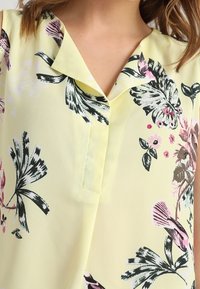 Blusa gialla con colletto, caratterizzata da motivi floreali in rosa, verde e nero. Tessuto leggero e setoso con bottoni.