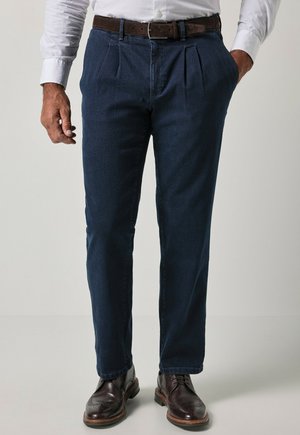 Mann trägt dunkelblaue plissierte Hose, braunen Ledergürtel, weißes Hemd und dunkelbraune elegante Schuhe, steht mit Händen in den Taschen.