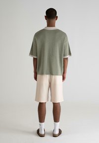Green textured short-sleeve shirt with a round neckline, paired with light beige shorts, worn with white socks and brown sandals.