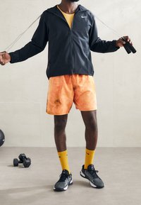 Chaqueta negra con cremallera, pantalones cortos naranjas con un patrón de tie-dye, calcetines amarillos y zapatillas deportivas negras. Sosteniendo una cuerda para saltar.