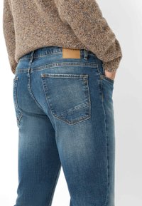 Blauwe spijkerbroek met een licht vervaagde was, twee achterzakken en een bruine leren patch op de tailleband. Textiel gebreide trui zichtbaar erboven.