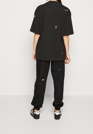 Henkilö pukeutuneena mustaan ylipitkään t-paitaan ja mustiin jogger-housuihin, joissa on valkoiset logot, seisoo valkoista seinää vasten, kädet rentoina sivuilla.