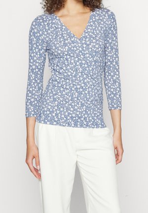 Femme portant un haut à manches longues bleu et blanc à fleurs avec col en V et un pantalon blanc taille haute, debout devant un fond uni.
