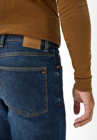 Jeans in denim blu scuro con cuciture arancioni a contrasto, dotati di una patch in pelle sulla vita e una mano visibile a sinistra.