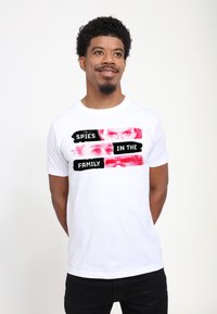 Camiseta de algodón blanca con un diseño gráfico que presenta un texto en rojo y negro "ESPÍAS EN LA FAMILIA" superpuesto sobre imágenes de dos ojos.