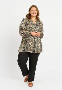 Locker sitzende Bluse in creme- und schwarzblumigem Muster mit V-Ausschnitt; kombiniert mit schwarzen Hosen und braunen Rutsch-Sandalen. Glatte Stofftextur.