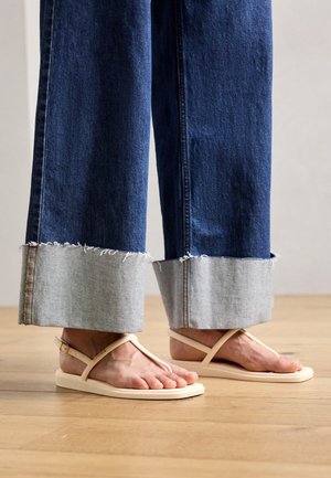 Pantalons larges en denim foncé avec des ourlets en denim clair effilochés, associés à des tongs beige avec un design en T et des semelles plates.