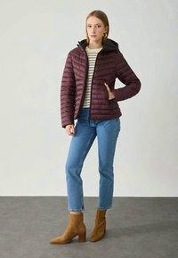 Giacca a vento bordeaux con cappuccio, motivo trapuntato, abbinata a una maglietta a righe, jeans blu e stivaletti beige.