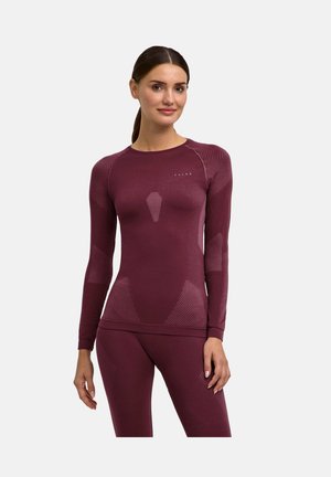 Kvinde med mørkt hår i hestehale iført en tætsiddende, langærmet, vinrød træningstop og matchende leggings mod en ensfarvet baggrund.