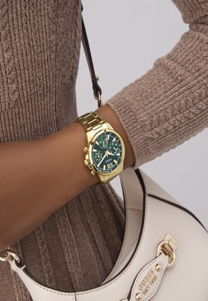 Montre-bracelet en or avec un cadran vert et des marqueurs horaires blancs sur un bracelet en métal doré. Haut beige avec une texture côtelée et un petit sac à main clair avec des accessoires.