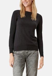 Schwarzer Pullover mit langen Ärmeln und Rundhalsausschnitt. Mit geripptem Saum und Bündchen. Weicher Stoff, enganliegende Silhouette. Kombiniert mit hellgrauen Jeans.