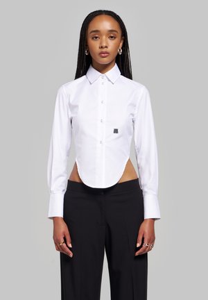 Femme aux longs cheveux tressés portant une chemise blanche boutonnée avec des côtés découpés et un pantalon noir, debout sur un fond gris uni.