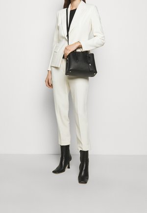 Costume blanc sur mesure avec pantalon slim, associé à des bottines noires à talons hauts. Un sac à main noir structuré est tenu dans une main.