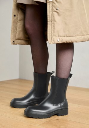 Čierne gumové členkové topánky s hladkým povrchom, elastickými bočnými panelmi a pevným podpätkom. Nosené s tmavými pančuchami a béžovým kabátom.