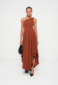 Καφέ φόρεμα με έναν ώμο, με εφαρμοστό κορσέ, ρέουσα ασύμμετρη άκρη και υφή υφάσματος. Συνδυασμένο με μαύρη τσάντα και ψηλές γόβες.