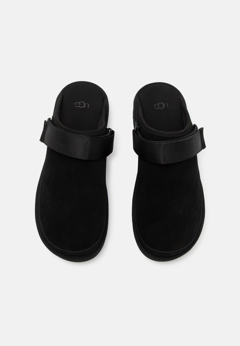 Sandali slip-on in morbida pelle scamosciata nera con una larga cintura regolabile. Texture soffice, forma arrotondata e fondello sagomato per il comfort. Design semplice e moderno.