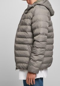 Sidan av en person som bär en grå quiltad pufferjacka med huva, vit skjorta och blå jeans mot en enfärgad bakgrund.