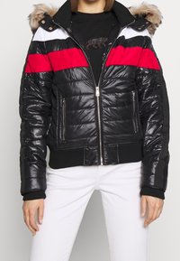 Puffjacka med svarta, röda och vita paneler, med en faux fur-hette, dragkedja fram och ribbade ärmslut och fåll. Texturerad, glansig yta.