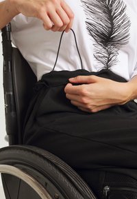 Osoba na wózku inwalidzkim poprawiająca czarne spodnie ze sznurkiem, ubrana w białą koszulę z czarnym wzorem piór.