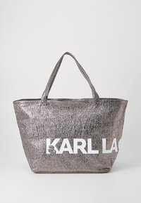 Metalinis sidabrinis pirkinių krepšys su struktūrizuota danga, turintis dvi rankenas ir didelį baltą "KARL LAGERFELD" logotipą priekyje.