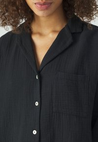 Schwarze Hemdbluse aus leichtem Strukturstoff, mit Kragen, Brusttasche und kontrastierenden cremefarbenen Knöpfen.
