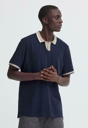 Ung mann i marineblå poloshirt med beige krage og ermekant, stående med sammenlignede hender og ser mot høyre mot ensfarget bakgrunn.