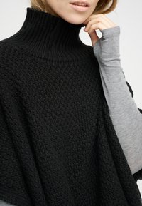 Svart stickad poncho med hög krage och strukturerat mönster, kombinerad med en ljusgrå långärmad tröja under.