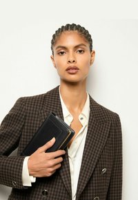 Fekete tárca arany lánc díszítéssel, kézben tartva. A modell barna kockás blazer-t visel csúcsos hajtókával egy fehér ing felett. Simább anyagok.