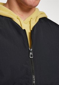 Μαύρο, ελαφρύ εξωτερικό μπουφάν με φερμουάρ πάνω από μια κίτρινη φλις κουκούλα. Διαθέτει λεία υφή και αντίθετα χρώματα στη γραμμή του λαιμού.