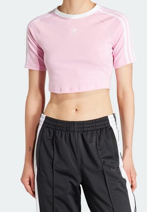 Personne portant un t-shirt court rose Adidas avec des rayures blanches sur les manches et un pantalon de survêtement Adidas noir avec des rayures blanches.