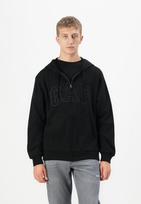 Young man wearing a black GAP zip-up hoodie and light gray jeans standing against a plain white background.