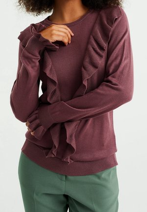 Femme portant un pull mauve avec un bord à volants sur le devant et aux poignets, associée à un pantalon vert, debout devant un fond uni.