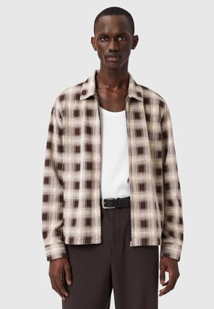 Chemise à carreaux marron et beige avec col, fermeture à boutons et manches longues, superposée à un débardeur blanc, associée à un pantalon foncé.