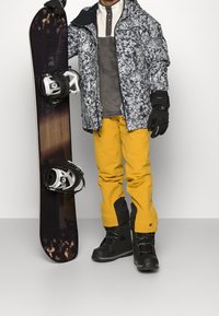 Snowboardista v bundě s květinovým vzorem, šedé střední vrstvě, žlutých kalhotách a černých botách, držící snowboard s bílými vázáními.
