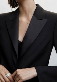 Musta bleiseri satiinikiilalikuksella, jossa on rakennetut hartiat ja pitkät hihat. Tasainen kangas ja istutettu malli syvällä pääntiellä.