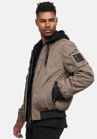 Giubbotto bomber in taupe con chiusura a zip, felpa nera sotto, tasche laterali e una toppa quadrata sulla manica sinistra. Tessuto testurizzato.