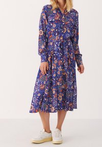 Sukienka z długim rękawem, zapinana na guziki, w niebieskim materiale z kwiatowym wzorem w kolorze czerwonym, pomarańczowym i białym. Zawiera pasek na sznurku i rozkloszowany dół. Połączona z białymi trampkami.