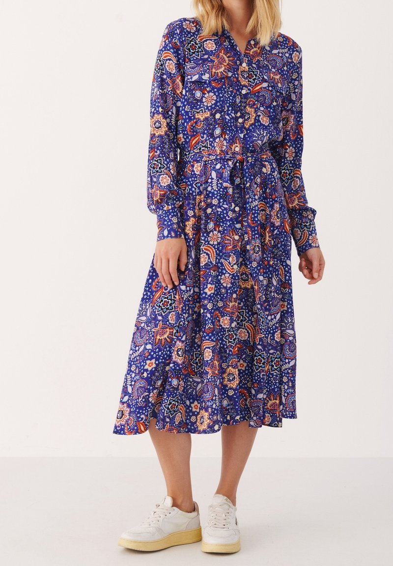 Sukienka z długim rękawem, zapinana na guziki, w niebieskim materiale z kwiatowym wzorem w kolorze czerwonym, pomarańczowym i białym. Zawiera pasek na sznurku i rozkloszowany dół. Połączona z białymi trampkami.