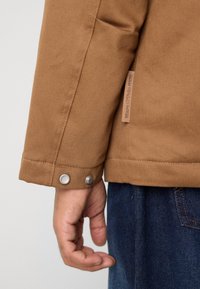 La mano de una persona cerca del puño de una chaqueta marrón con botones a presión metálicos y la etiqueta "Marc O'Polo Denim", vistiendo jeans de mezclilla azules.
