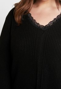 Suéter negro de punto con escote en V y borde de encaje negro. Textura acanalada con corte holgado, que muestra un diseño cómodo y casual.