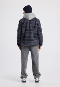 Blue plaid jacket with a gray hood, worn over gray jeans. Features ribbed navy beanie and white/gray sneakers with dark accents.